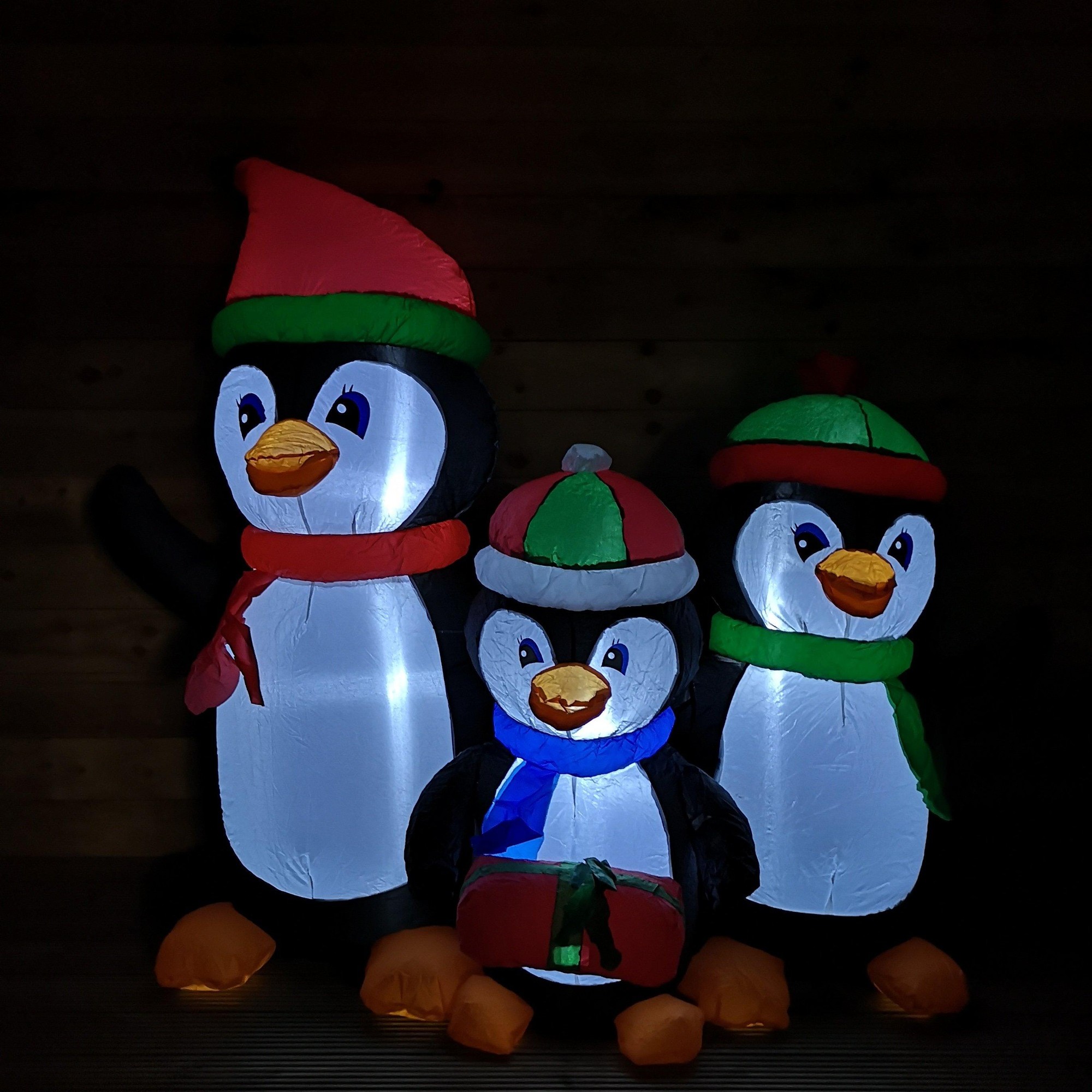 Three inflatable penguins in festive attire, featuring red and green hats, bright white LED lights, and colorful scarves, standing together against a dark background.