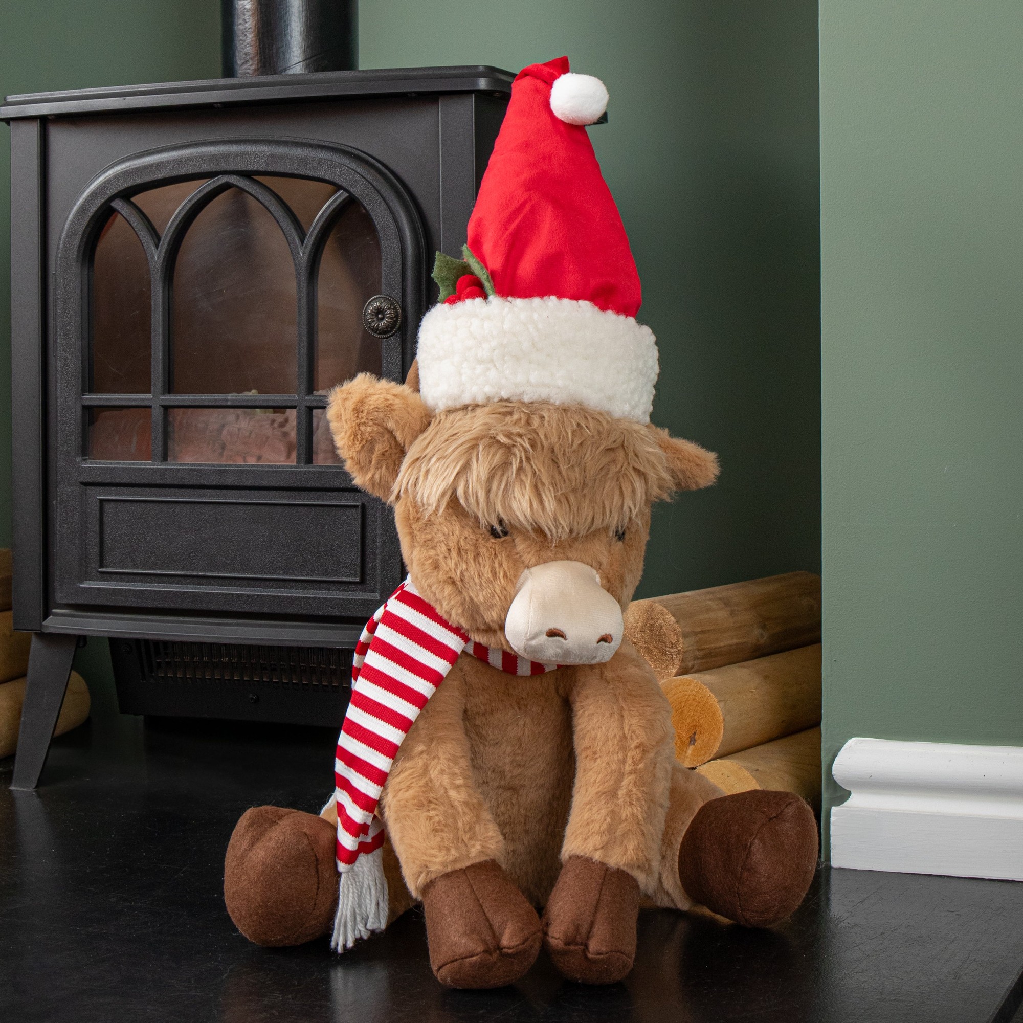 36cm tall plush sitting Highland boy cow wearing a red Santa hat and white fur trim, with a striped red and white scarf, seated beside a black stove.