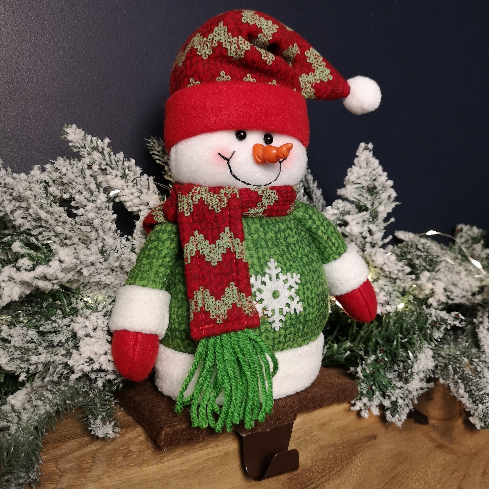 Plush Christmas snowman decoration with a green body, red hat, and scarf, featuring a white snowflake design, set against a snowy backdrop.