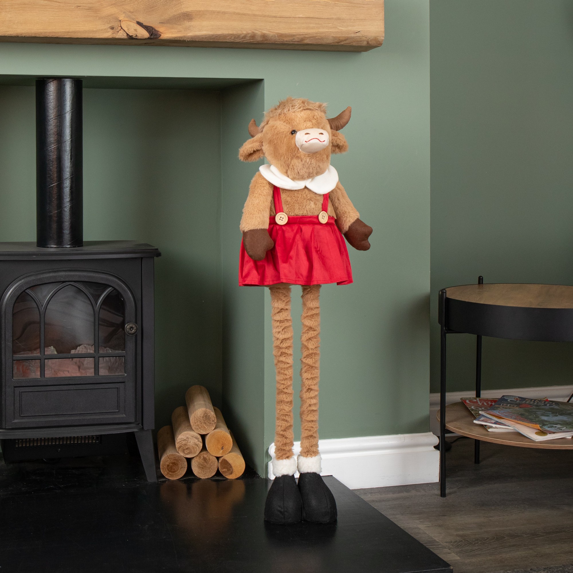 Tall plush Highland girl cow with extendable legs, wearing a red dress and white collar, brown fur, and black shoes, standing beside a cozy fireplace.