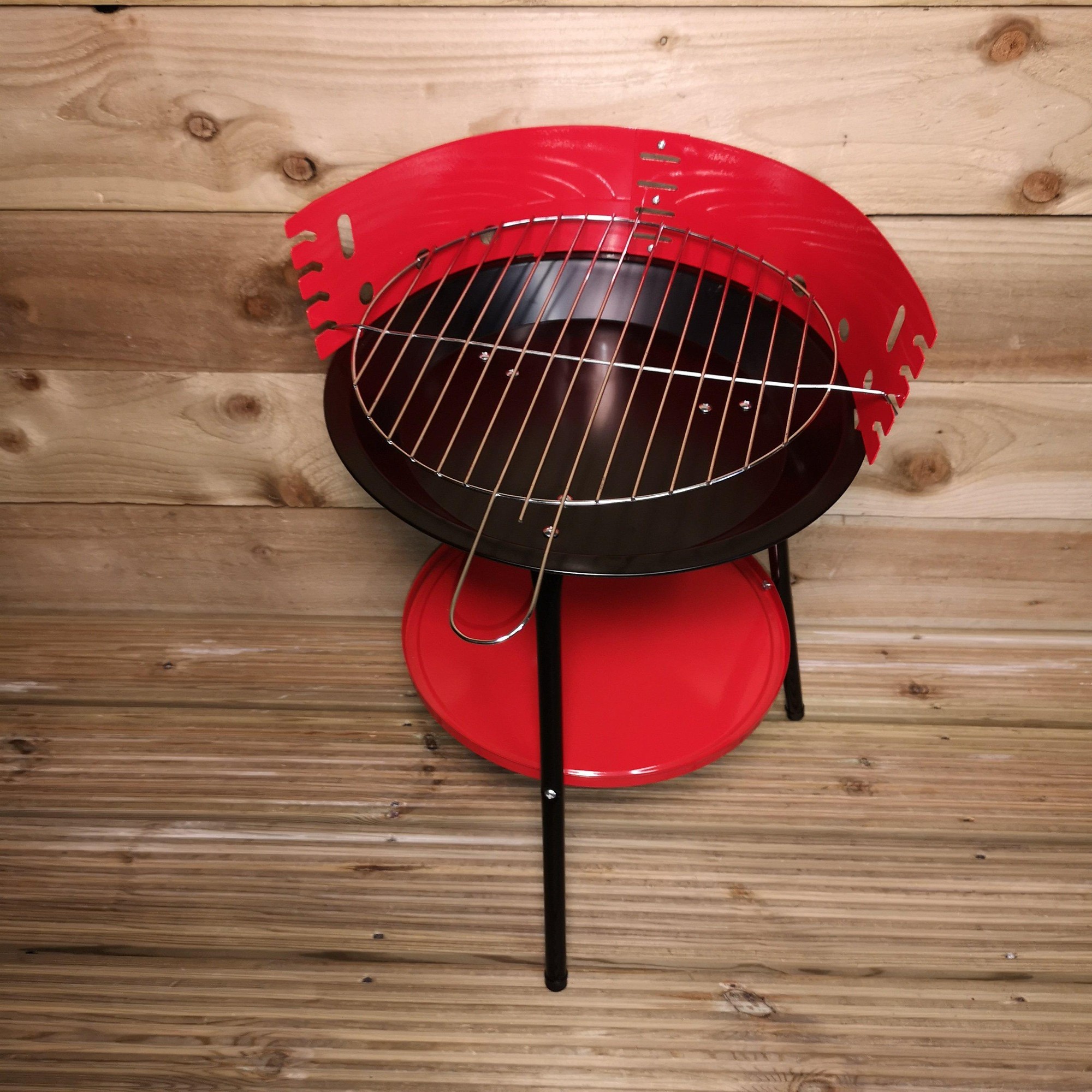 14-inch round garden barbecue in red with a metal wind shield, a chrome grill, and a lower shelf, set against a wooden backdrop.
