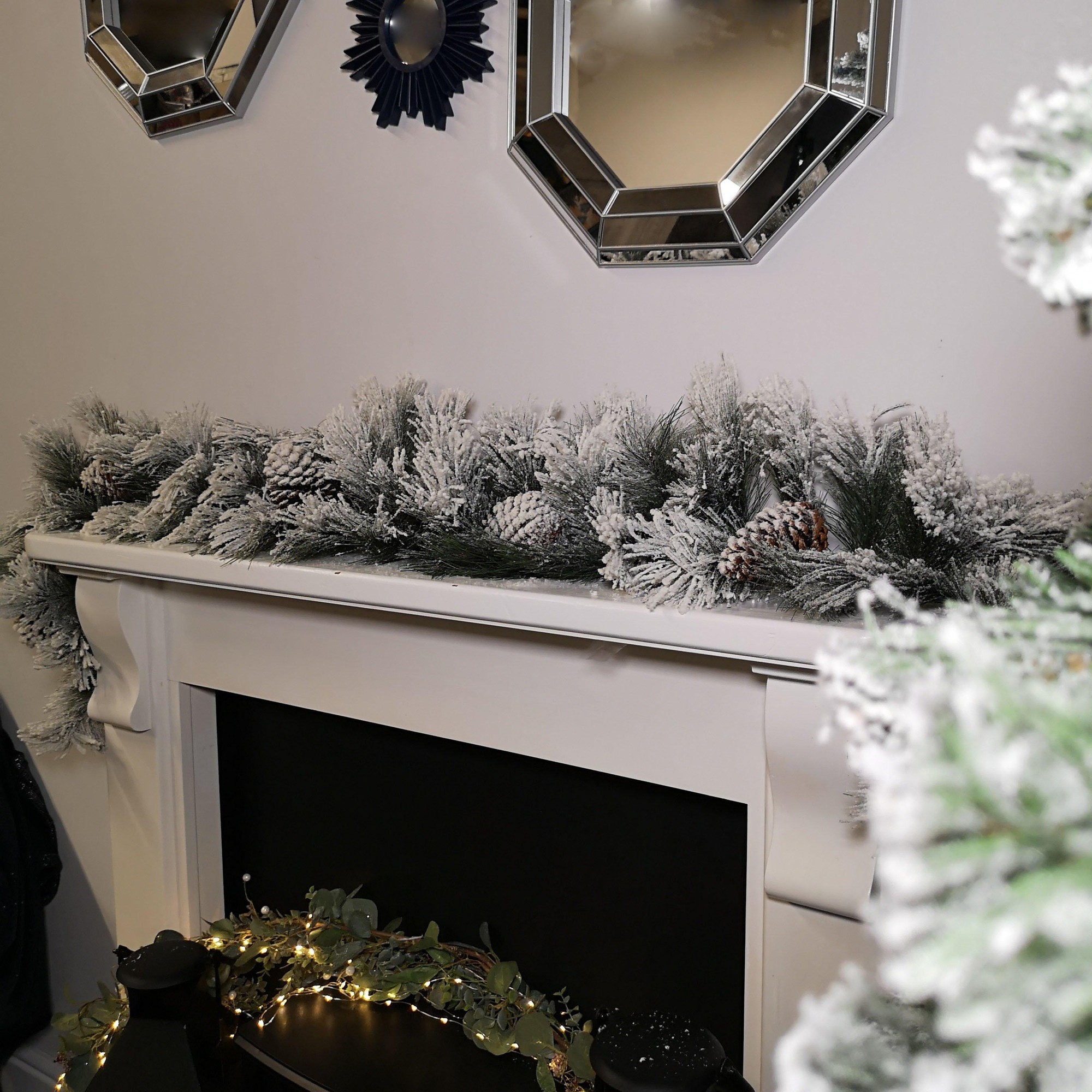 A 6ft snow-flocked green garland adorned with pine cones, featuring lush greenery and a frosted appearance, displayed on a white mantel with mirrors in the background.