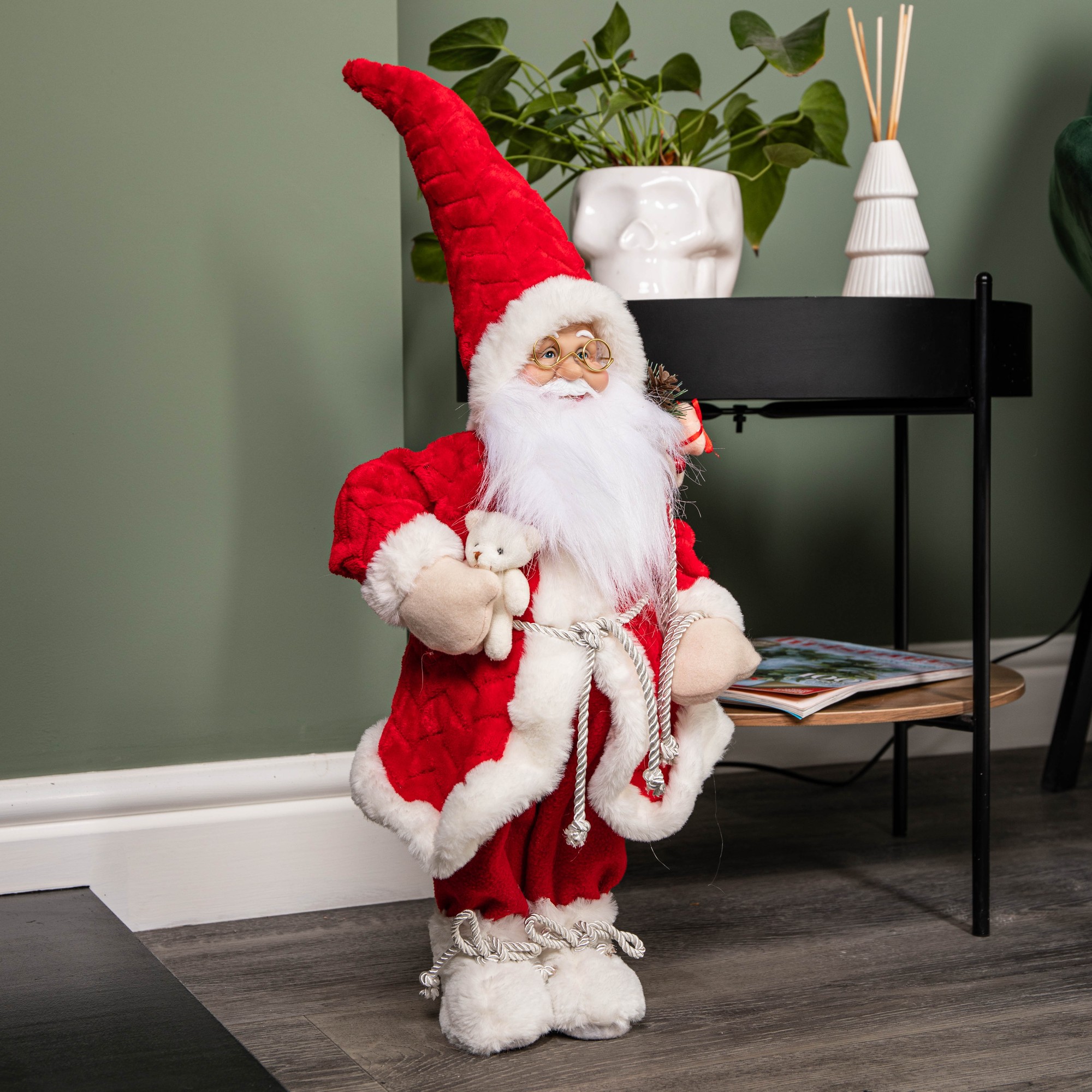 Large Santa Claus decoration in a red quilted coat and hat, holding a teddy bear, with white beard and boots, set against a green wall and modern decor.
