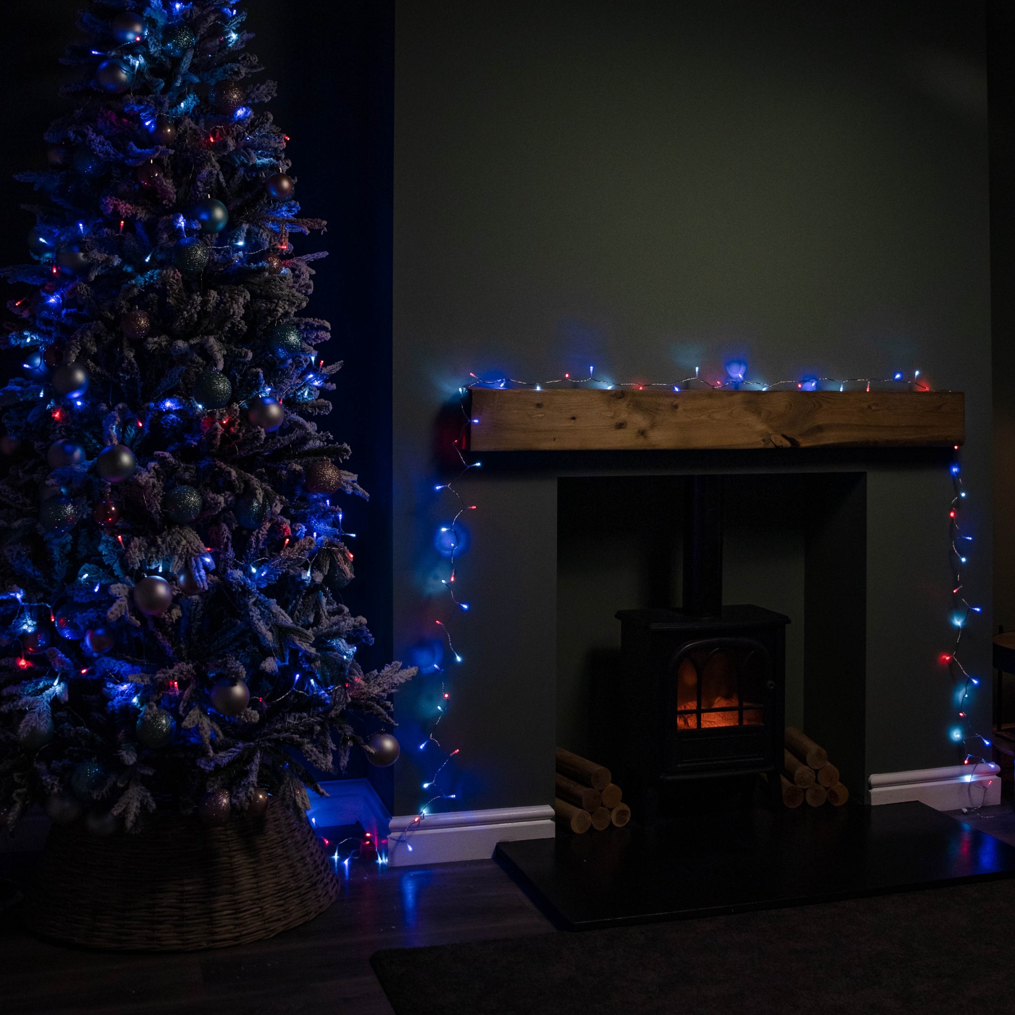 SupaBrights Christmas string lights in blue, red, and white on a clear cable, illuminating a decorated tree and mantelpiece beside a cozy fireplace.
