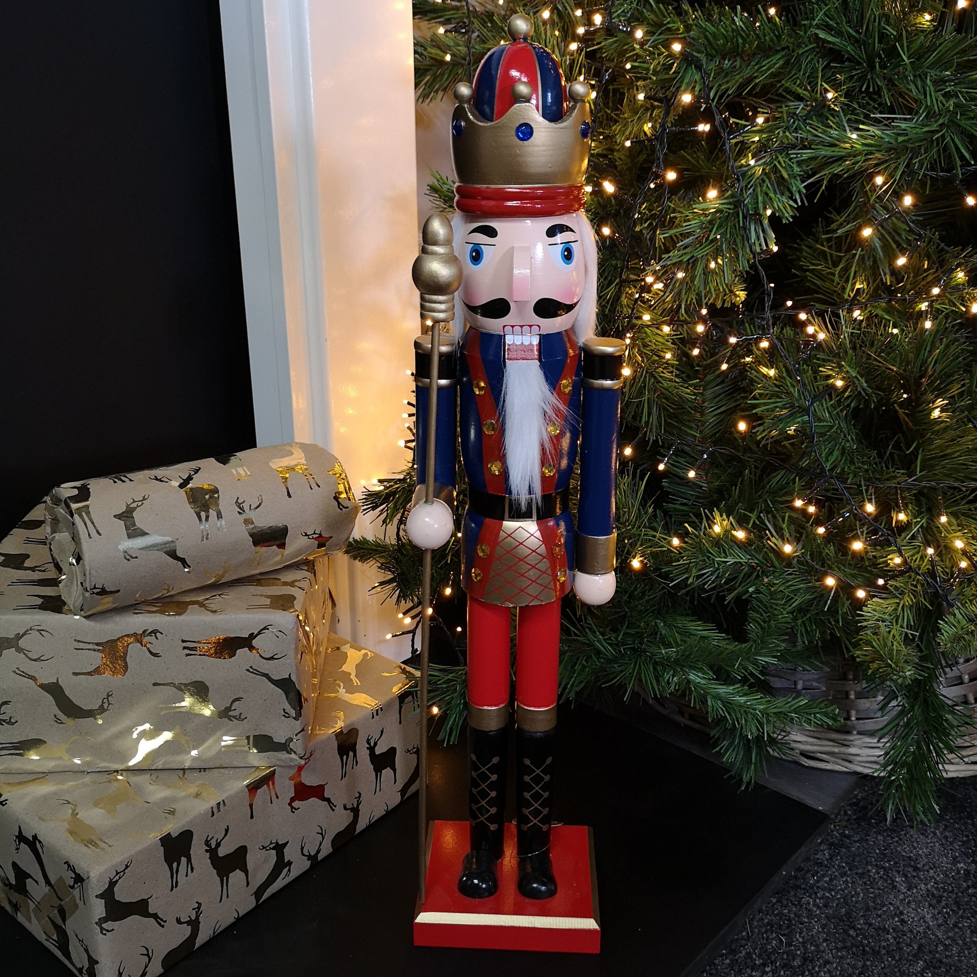 Traditional wooden nutcracker soldier decoration, 60cm tall, features a blue body, red pants, gold crown, and a white beard, standing beside wrapped gifts and a festive tree.