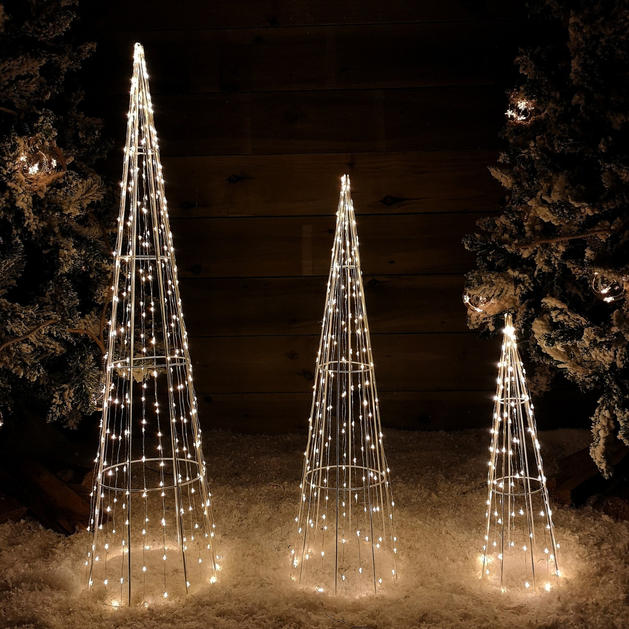 Three white cone-shaped LED trees in varying heights, adorned with 640 warm white lights, set against a dark wooden background and snowy ground.