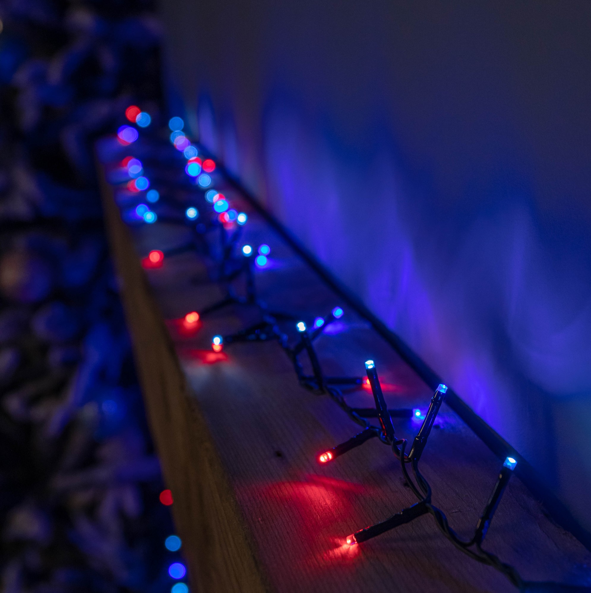 Colorful 240 LED Christmas string lights in blue, white, and red, featuring a timer, arranged along a wooden surface with a blurred festive background.