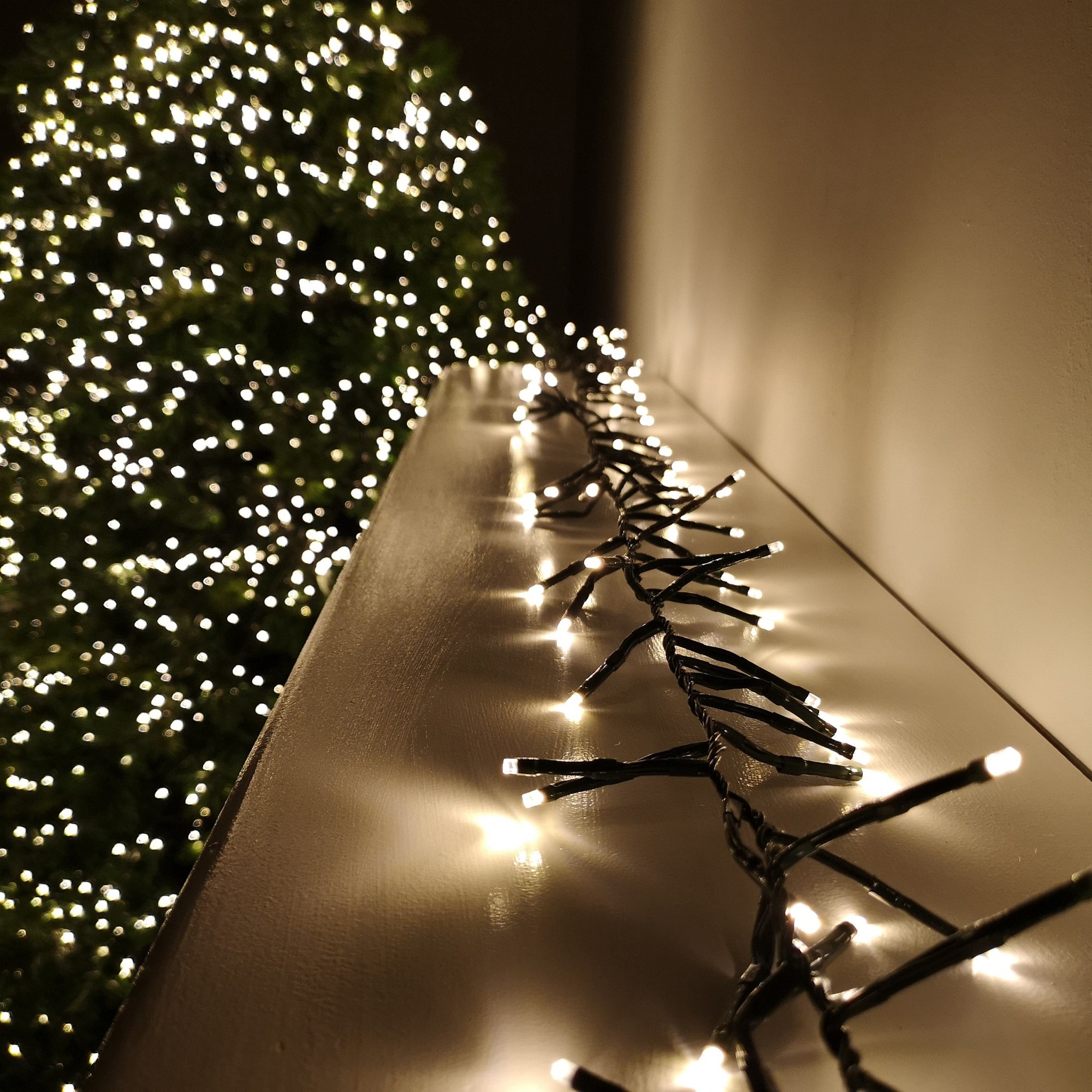 Warm white LED cluster Christmas lights on a dark green wire, featuring a timer, draped along a surface with a blurred green tree in the background.
