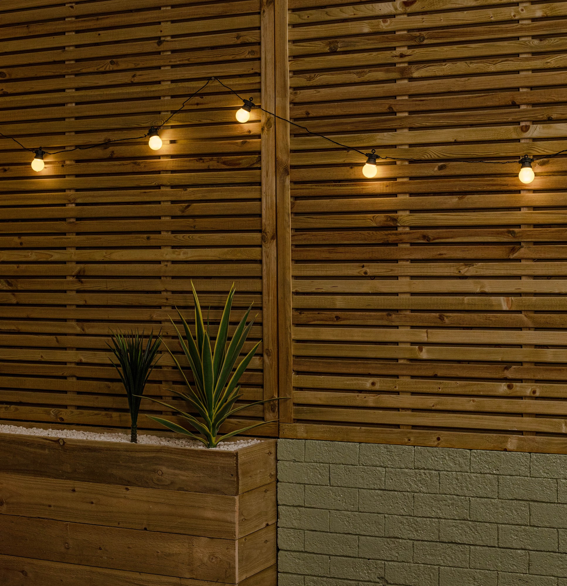 Connectable party festoon lights with warm white LEDs hang against a wooden slatted wall, illuminating a modern outdoor space with two potted plants below.