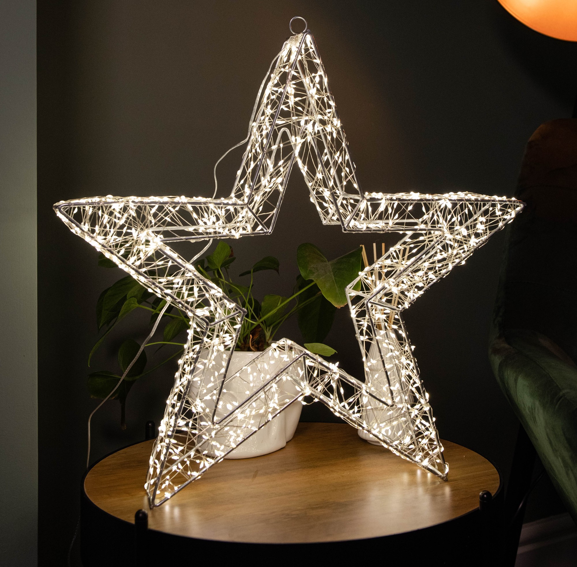 Silver dewdrop star decoration with 1440 warm white LEDs, featuring a timer function, set against a dark background with a plant and lamp nearby.