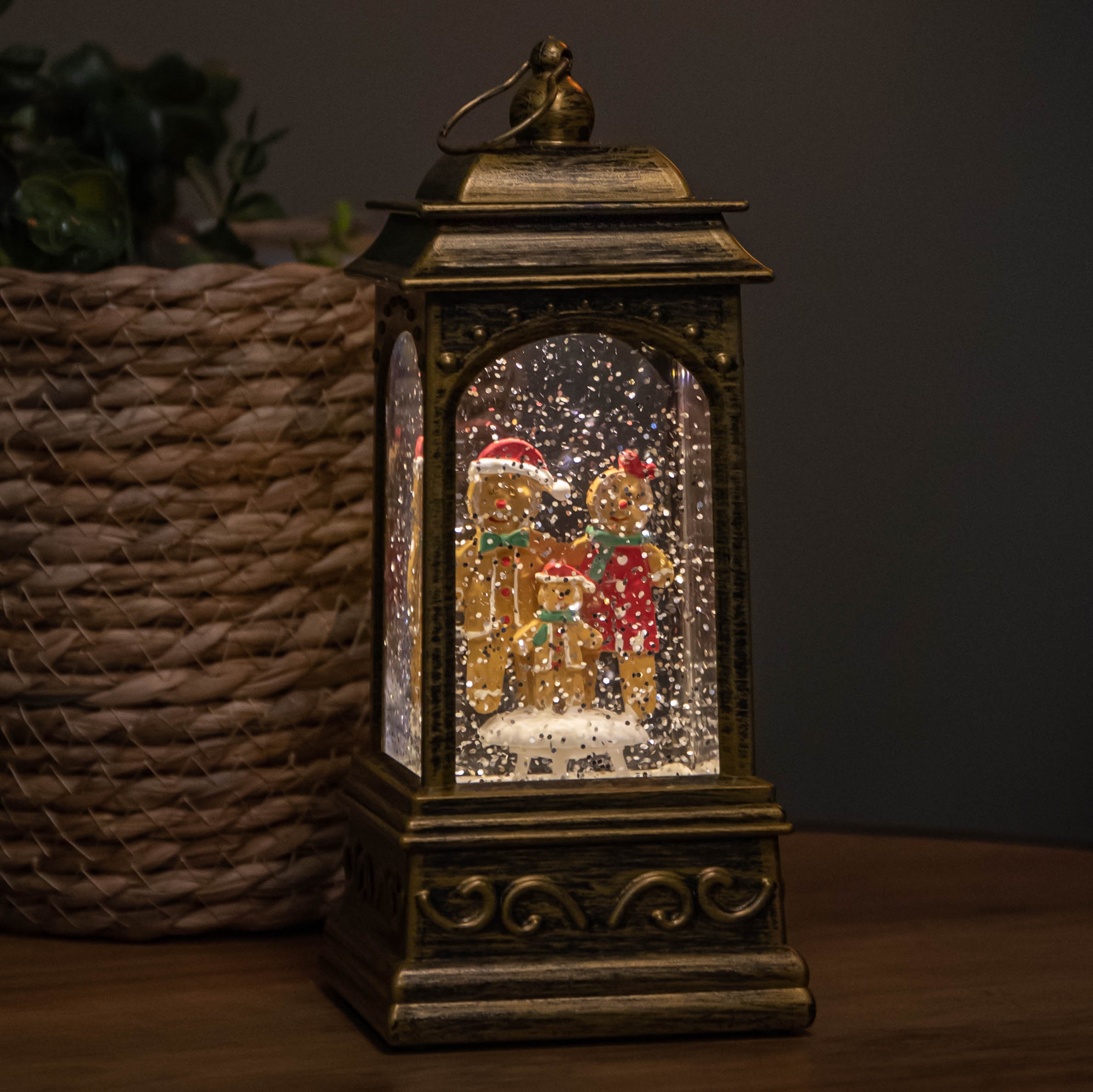 A decorative lantern featuring a gingerbread family, illuminated by LEDs, with a snow-like effect inside, set against a textured, gold-toned frame.