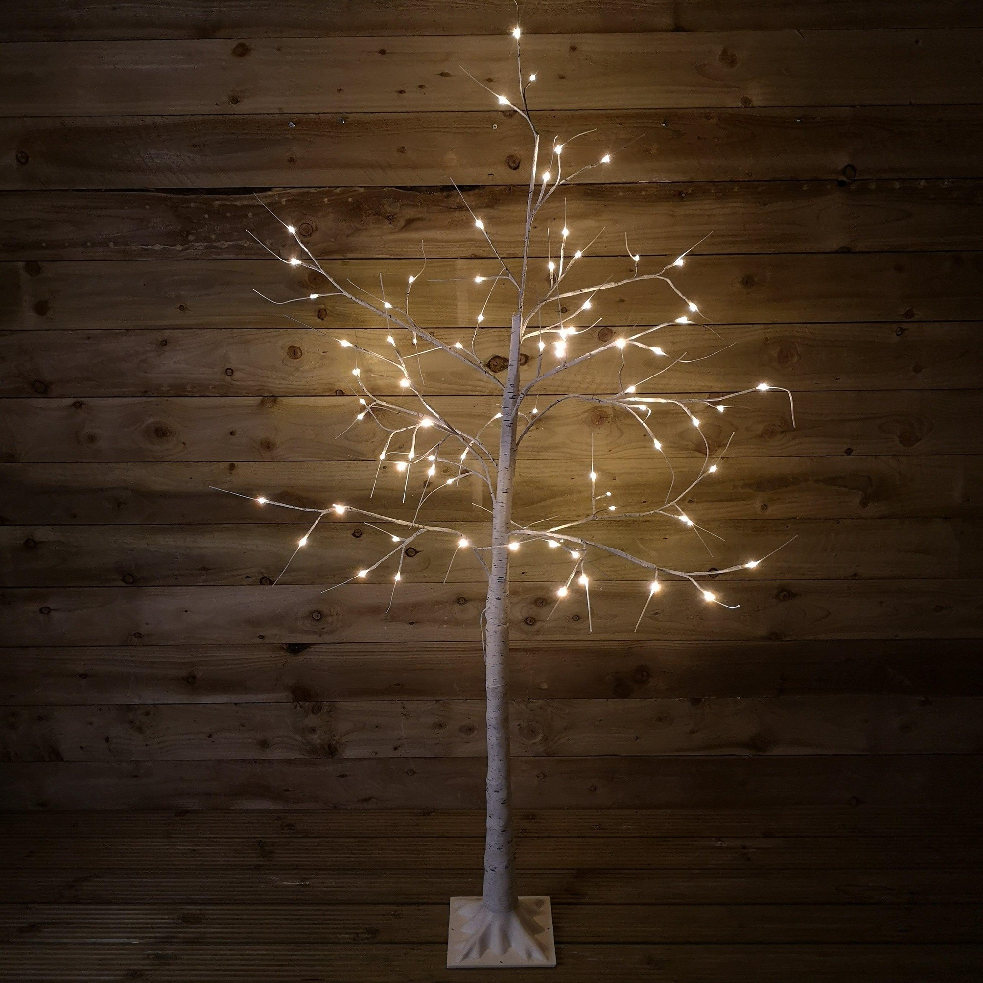 A 1.8M tall birch tree with a white trunk and branches, adorned with 80 warm white LED lights, standing on a square white base against a wooden backdrop.