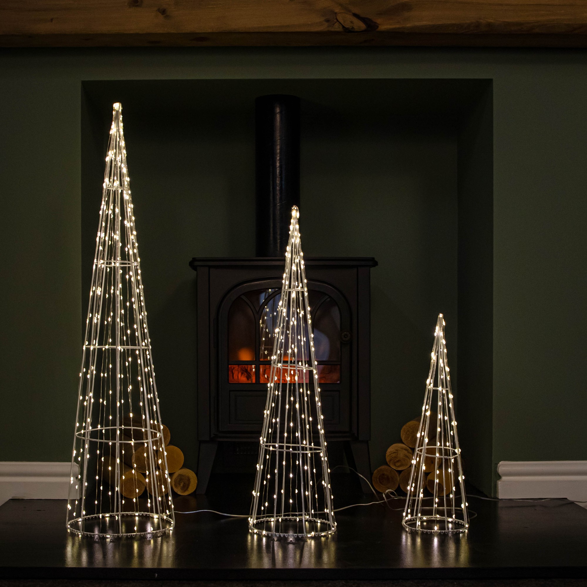 Set of three white metal dewdrop cone trees, varying in height, illuminated with warm white LEDs, placed in front of a black stove and green wall.