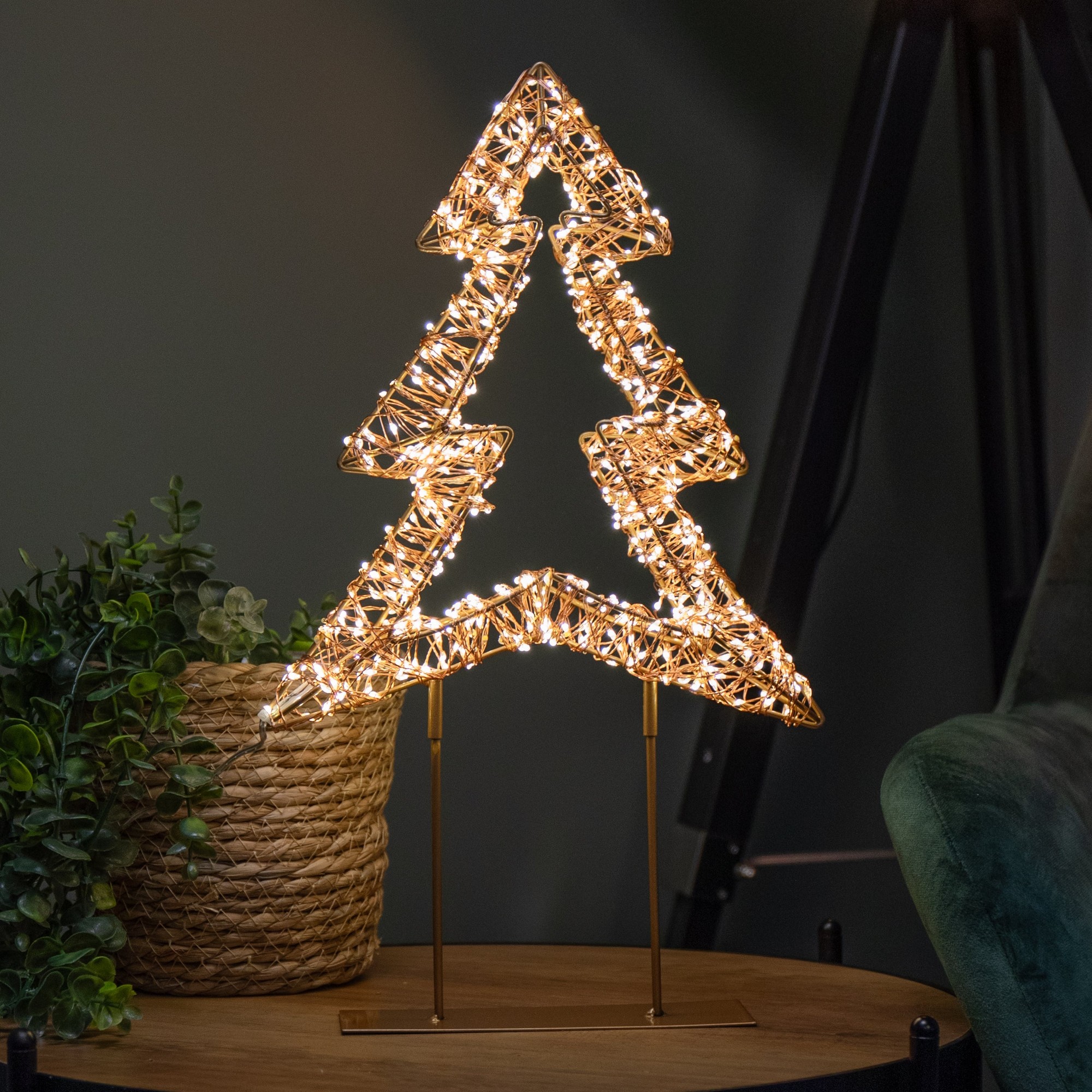 45cm rose gold Christmas tree made of metal with twinkling warm white LEDs, featuring a decorative design on a sturdy metal stand, placed beside a woven basket and green foliage.