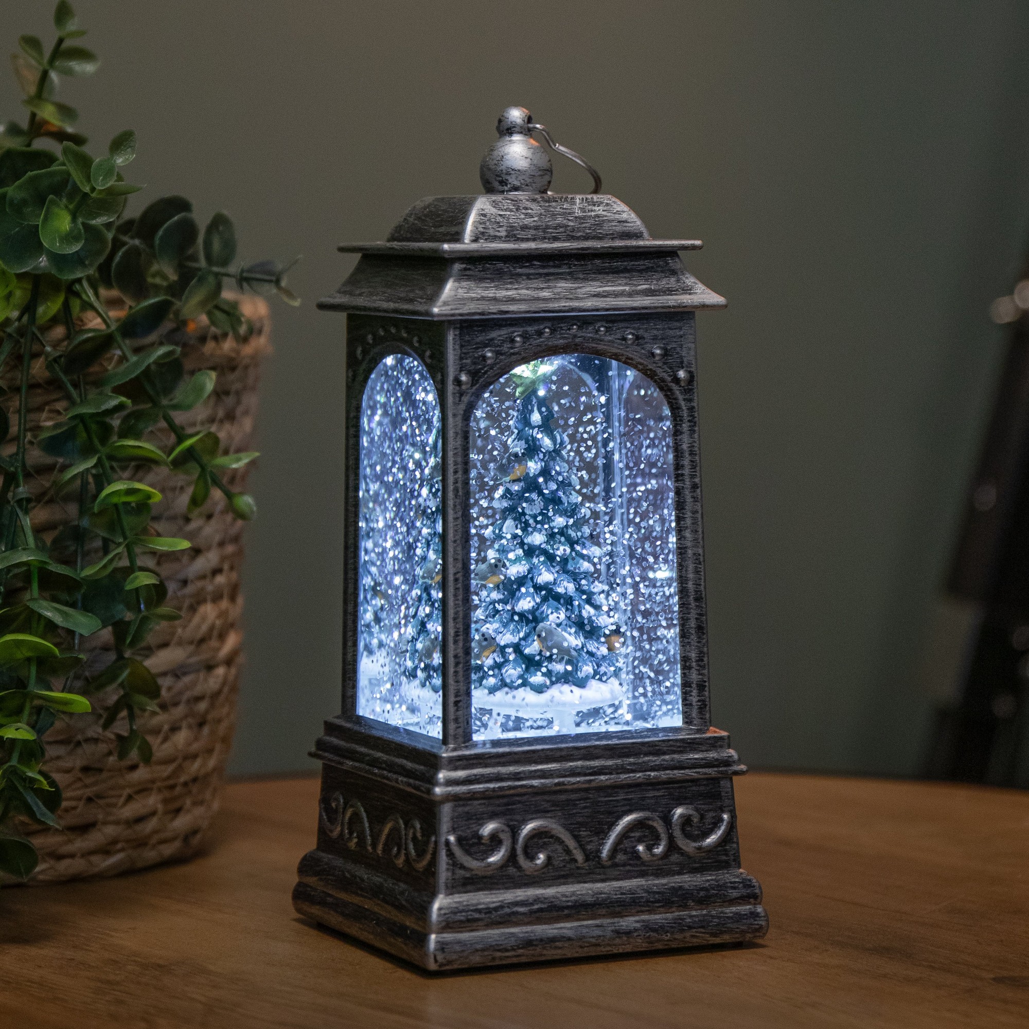 A 21cm silver water spinner lantern featuring a snowy Christmas tree and robins, illuminated by white LEDs, with a textured black base and clear glass panels.