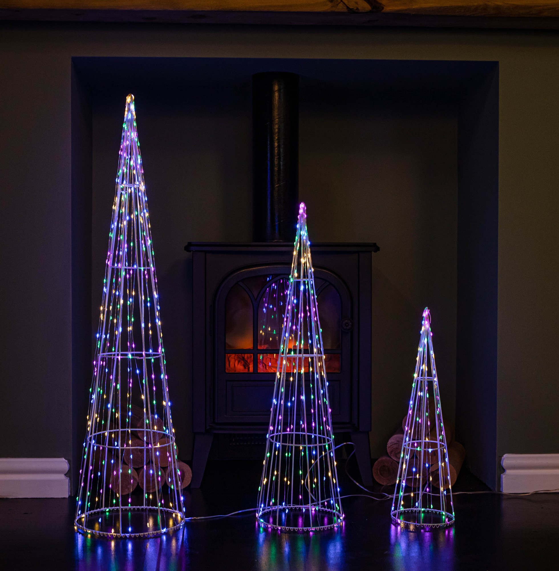Set of 3 light-up white metal dewdrop Christmas cone trees featuring multi-colored LEDs, displayed in front of a cozy fireplace.