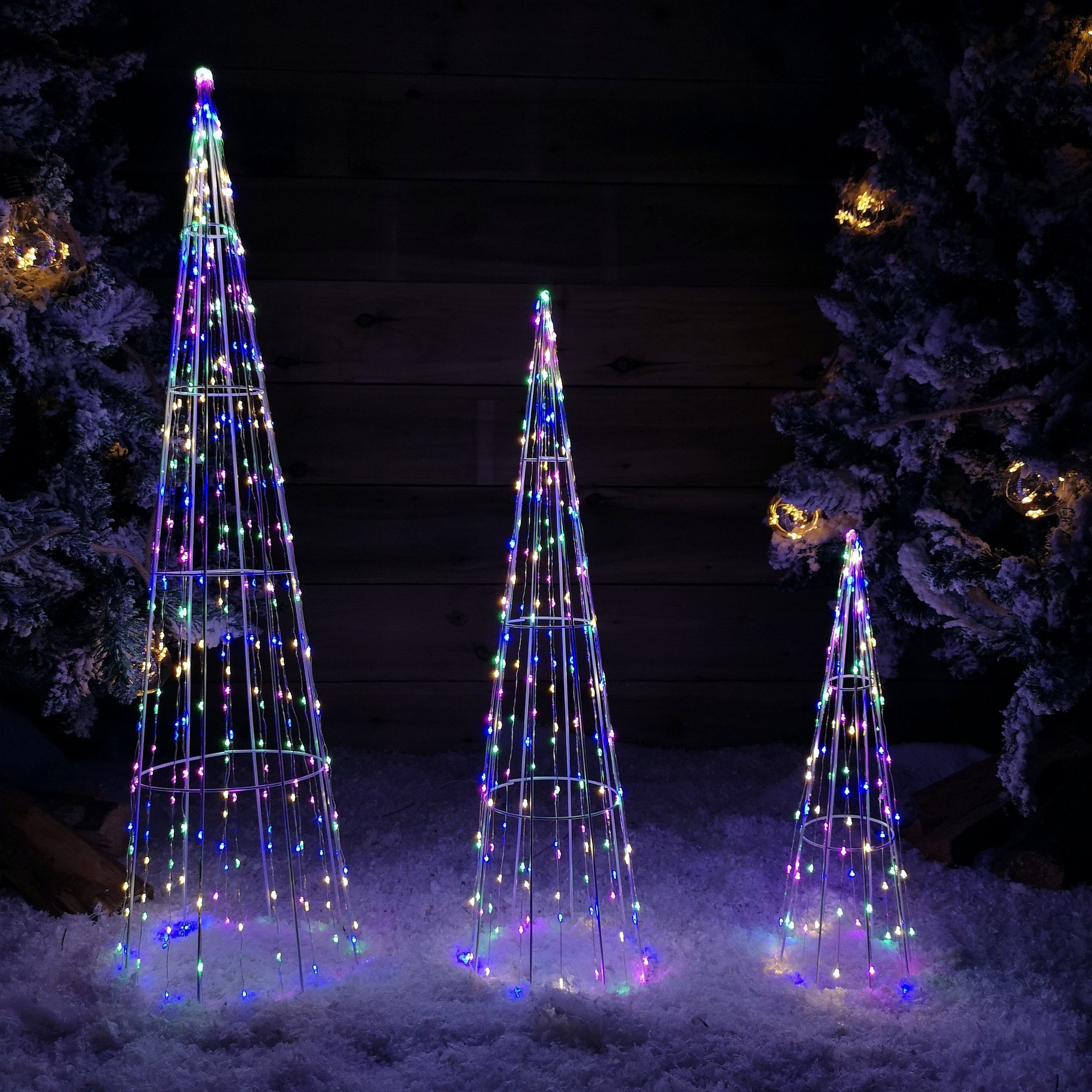 Three white cone-shaped trees made of wire, adorned with 640 multi-colored LEDs, set against a snowy backdrop, creating a festive ambiance.
