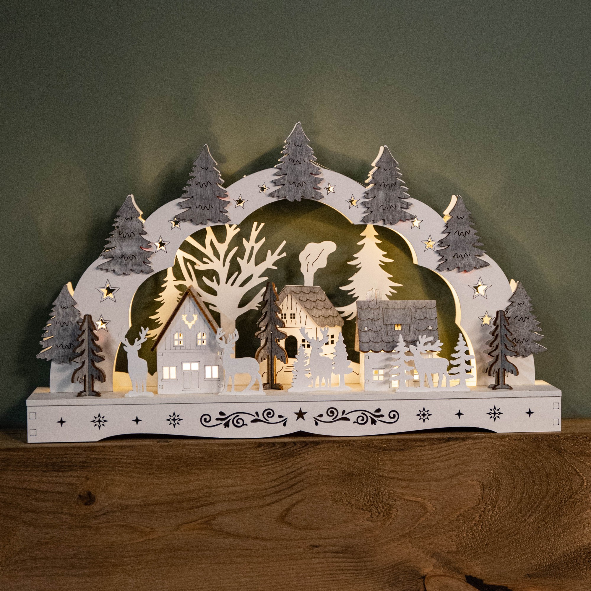 Battery-operated white wooden reindeer and village scene candle bridge features LED lights, intricate cut-out trees, and houses with grey roofs, set against a green background.