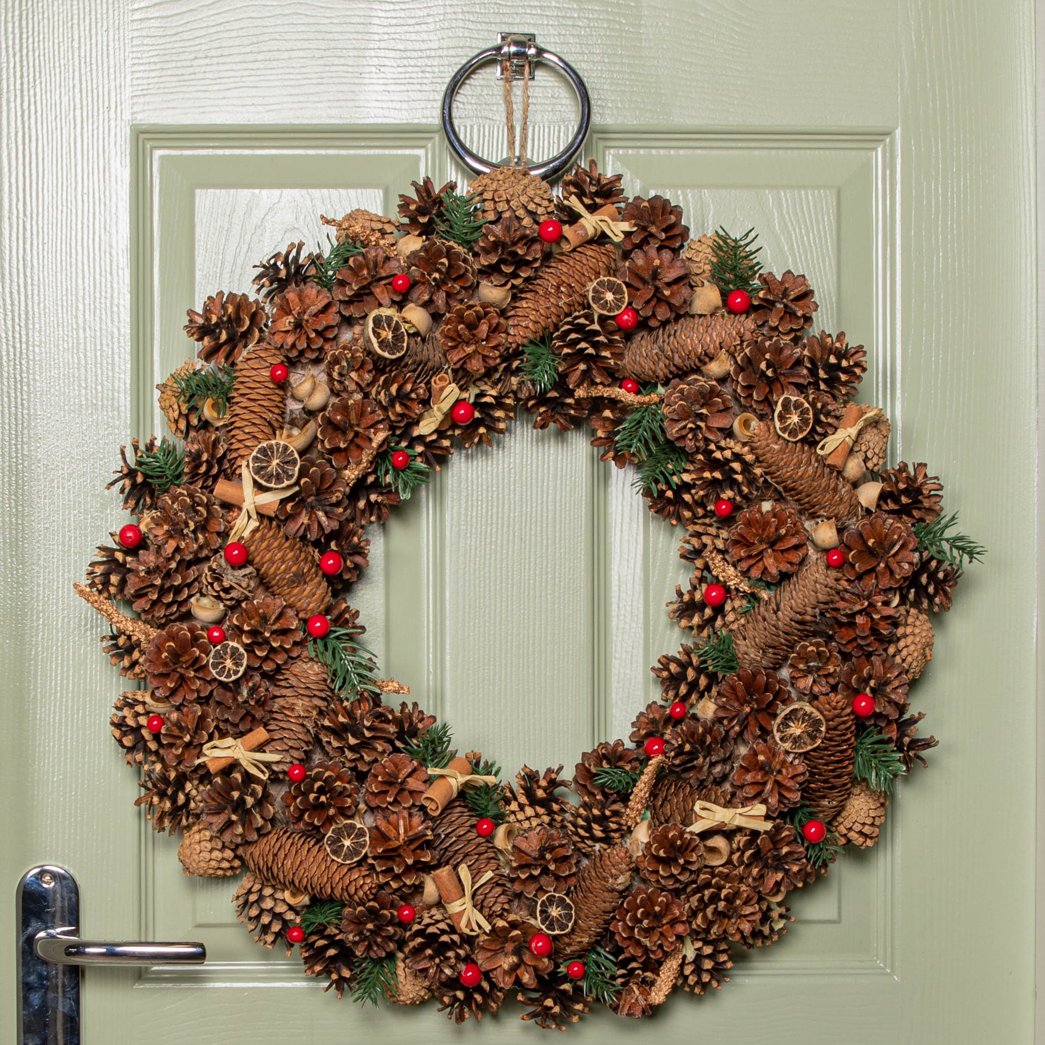 A 60cm circular Christmas wreath made of pine cones, red berries, and dried fruit, adorned with natural twine and decorative elements, hanging on a green door.