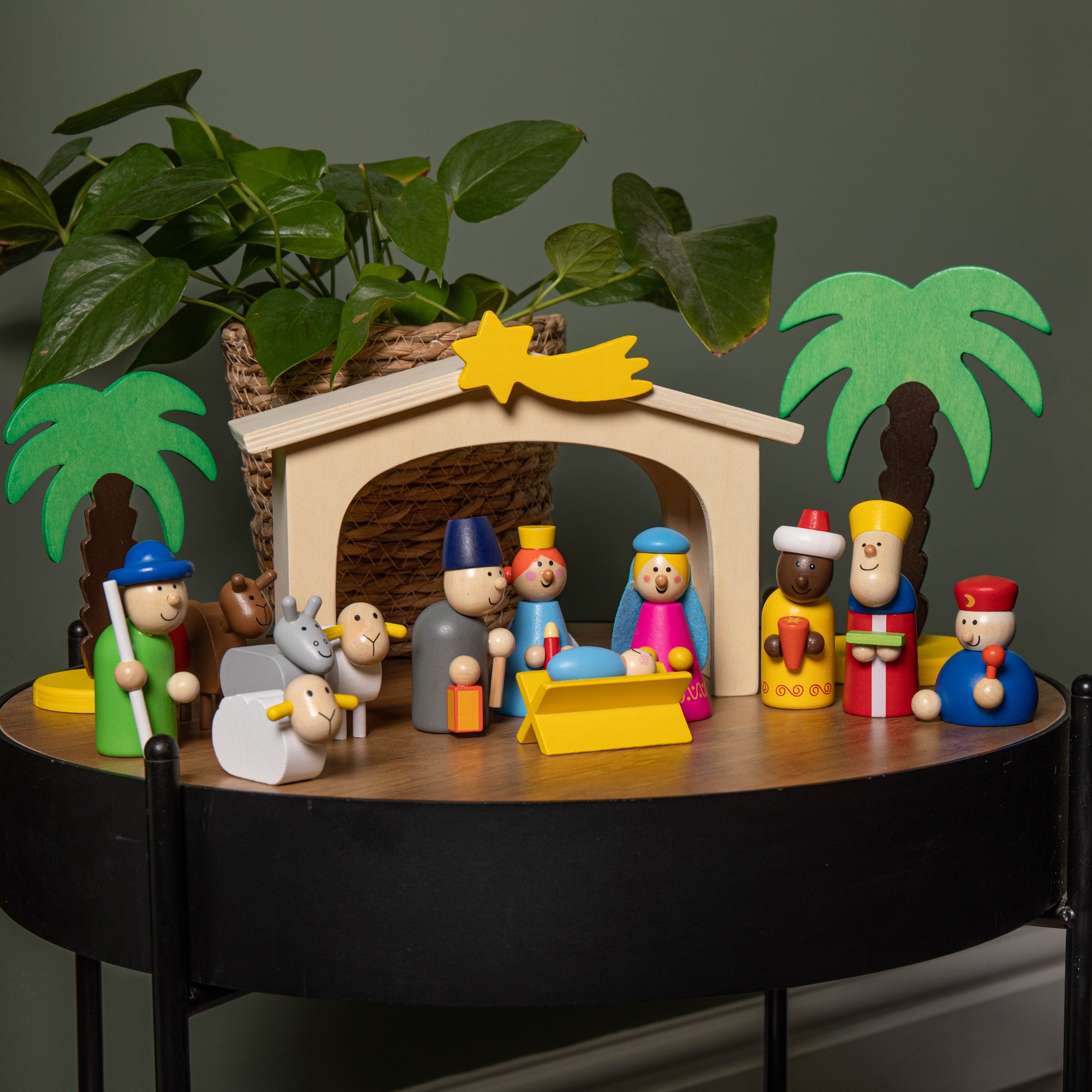 Wooden Christmas nativity scene playset featuring multi-colored figures, including Mary, Joseph, animals, and palm trees, set under a yellow star, on a round table.