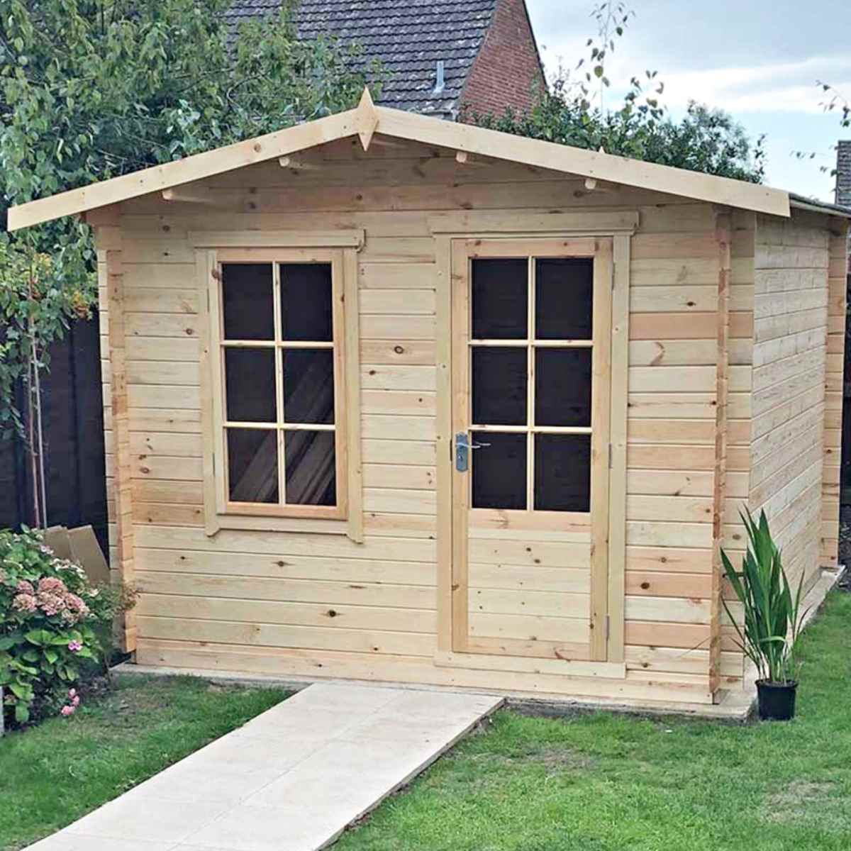 Light wood log cabin summerhouse with a sloped roof, double front doors, and two windows, set in a garden with greenery and a stone pathway.