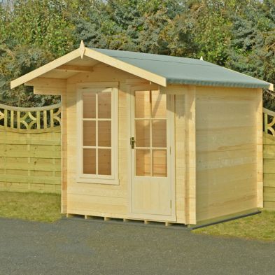 A wooden log cabin summerhouse with a light wood finish, featuring a sloped grey roof, double front doors, and large windows, set against a green fence and trees.