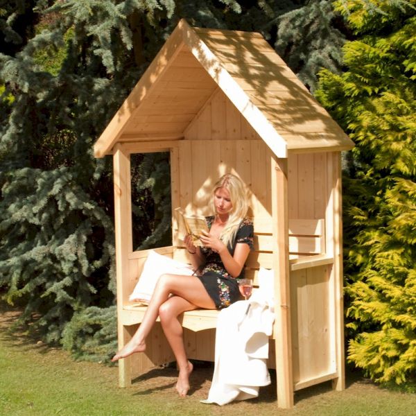 Natural wood garden arbour seat with a slanted roof, featuring a spacious bench and a woman reading, surrounded by lush greenery.