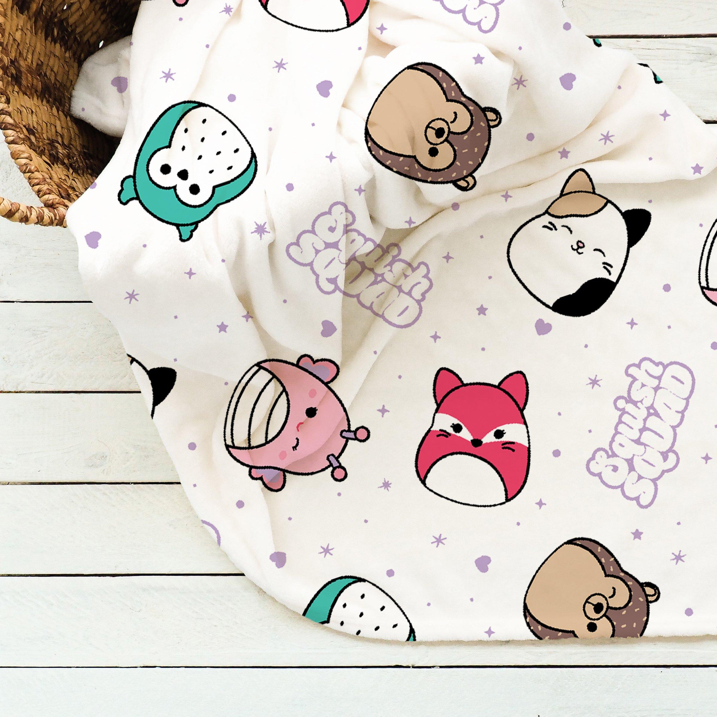 Soft fleece blanket featuring a playful pattern of colorful Squishmallows characters on a light background, with shades of pink, green, and brown.