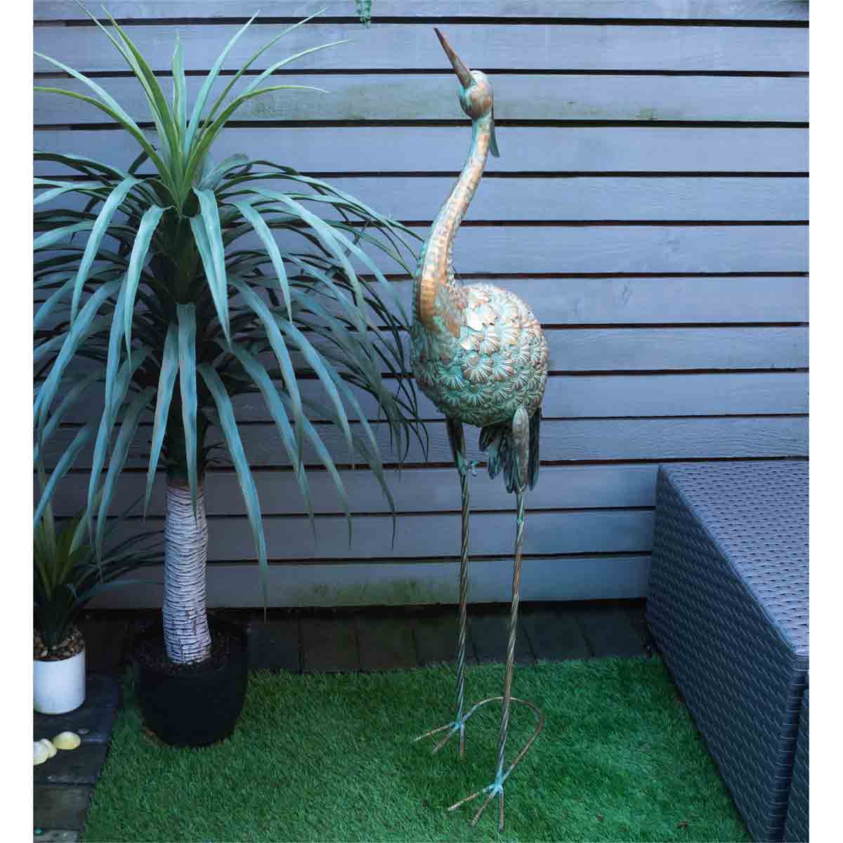 A 104cm tall gold metal heron garden ornament with intricate detailing, standing on slender legs beside a potted plant and a textured outdoor sofa.