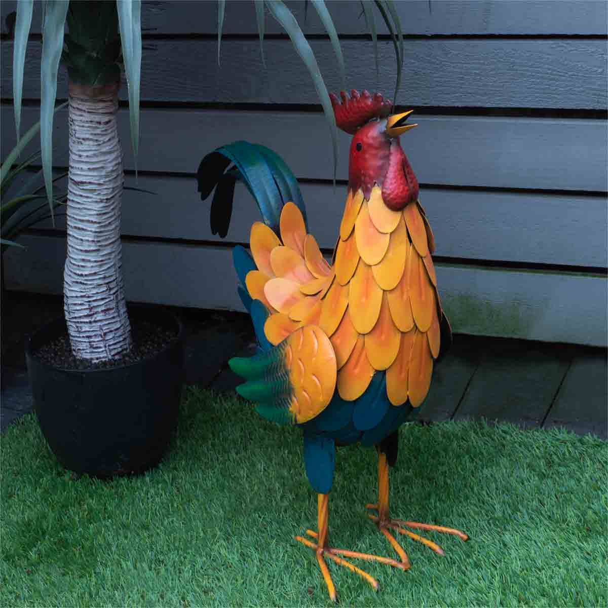 Colorful metal cockerel garden ornament, 46cm tall, featuring vibrant orange, yellow, green, and red feathers, with a detailed beak and tail, set against a grassy background.