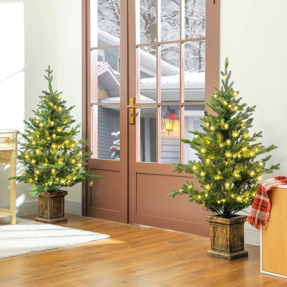 Two 4ft artificial Christmas trees in green with realistic tips and warm white lights, placed in decorative pots, flanking a doorway with a winter scene visible outside.
