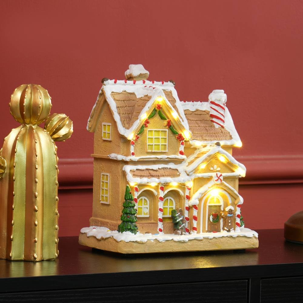 Lighted and musical Christmas candy house in brown with white icing, featuring a gingerbread man, candy canes, and a decorated door, set against a red wall.