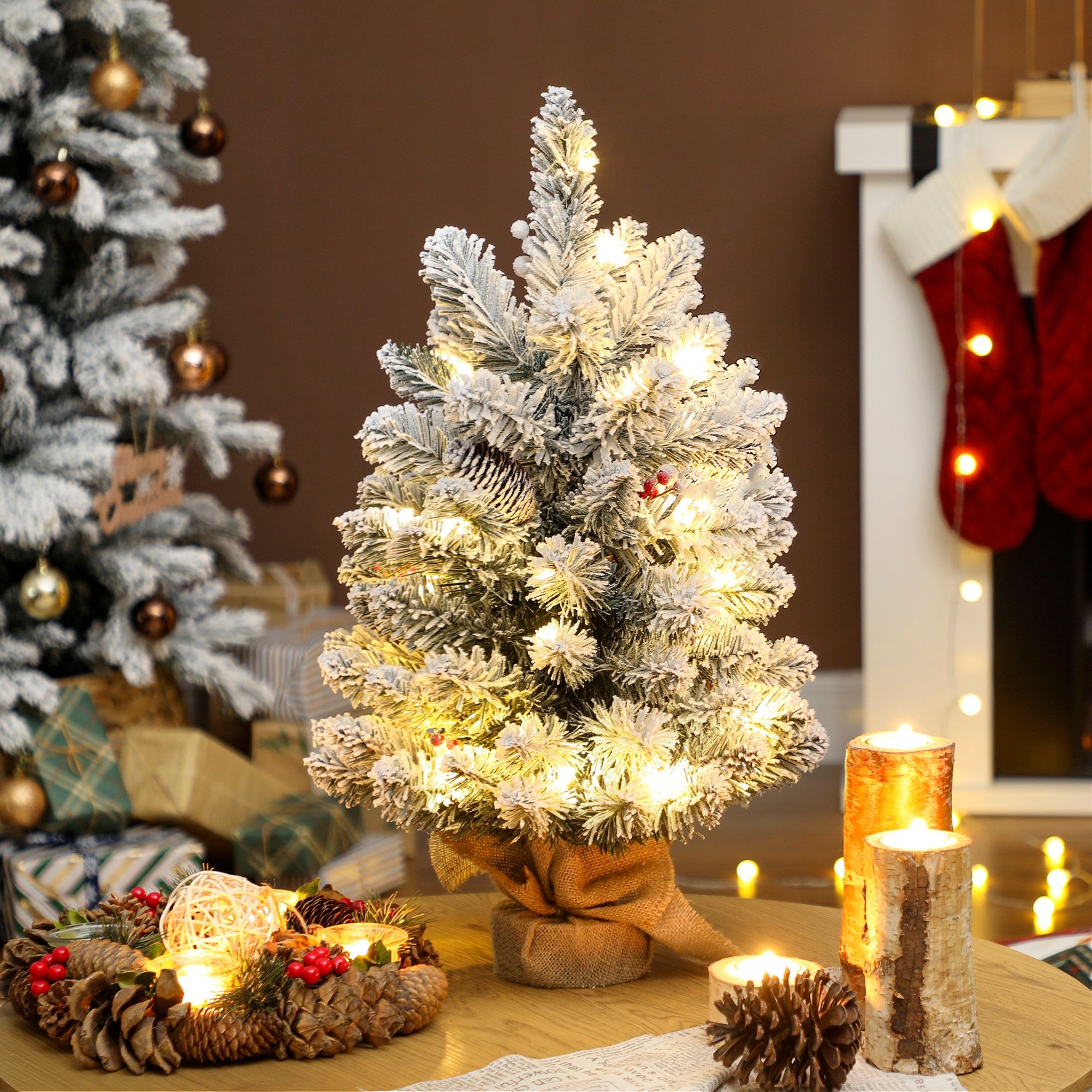 Small, 2ft green Christmas tree with snow-flocked branches, pre-lit with warm white lights, placed on a wooden table, surrounded by holiday decorations and candles.