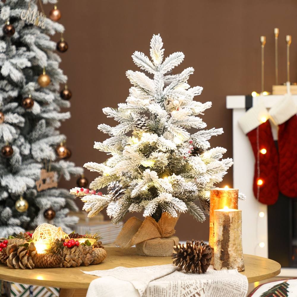 60 cm snow-flocked green tabletop Christmas tree with warm white lights, adorned with a burlap base, surrounded by festive decor like candles and pinecones.