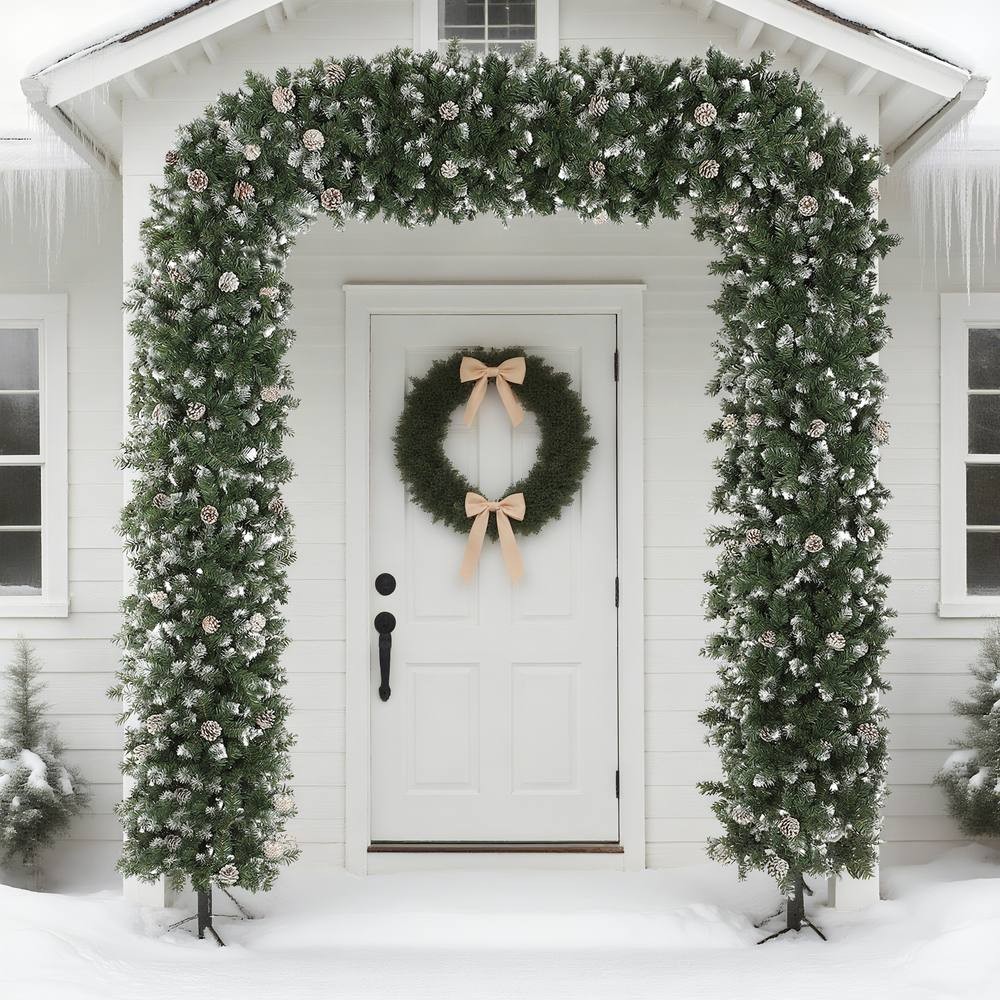 Snow-frosted green artificial Christmas tree arch adorned with pinecones, set against a white house with a wreath and a snowy entrance.