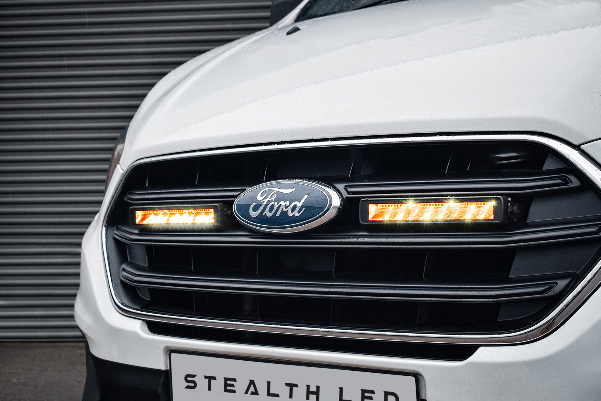 White Ford Transit Custom grille featuring a sleek black design with twin 10" flashing luminous LED lights, showcasing bright orange illumination and a prominent Ford logo.