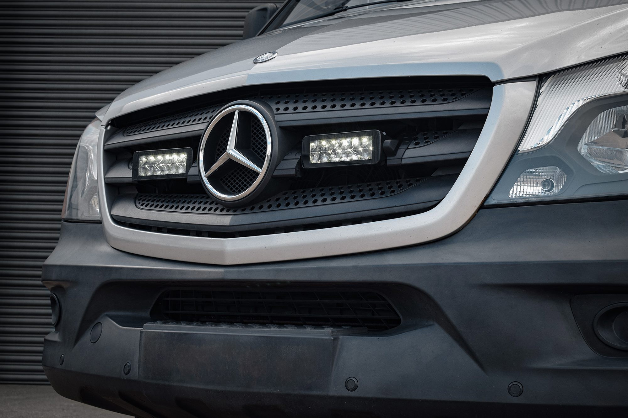 Twin 7" luminous LED lights integrated into a silver Mercedes Sprinter grille, showcasing a sleek design with a black mesh background and the iconic Mercedes emblem.