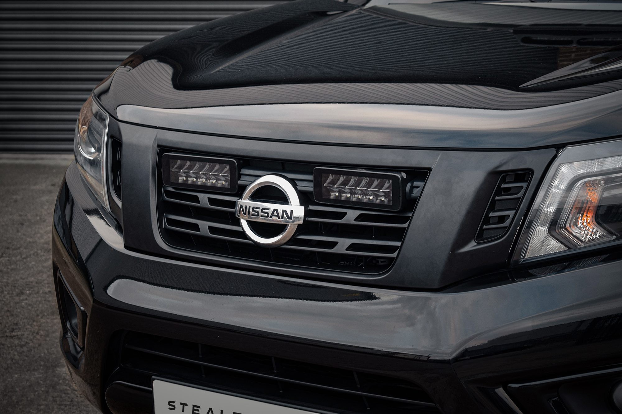 Black Nissan Navara D23 front grille featuring a chrome Nissan emblem, integrated with twin 7" luminous LED lights, showcasing a sleek design and high brightness of 10,080 lumens.
