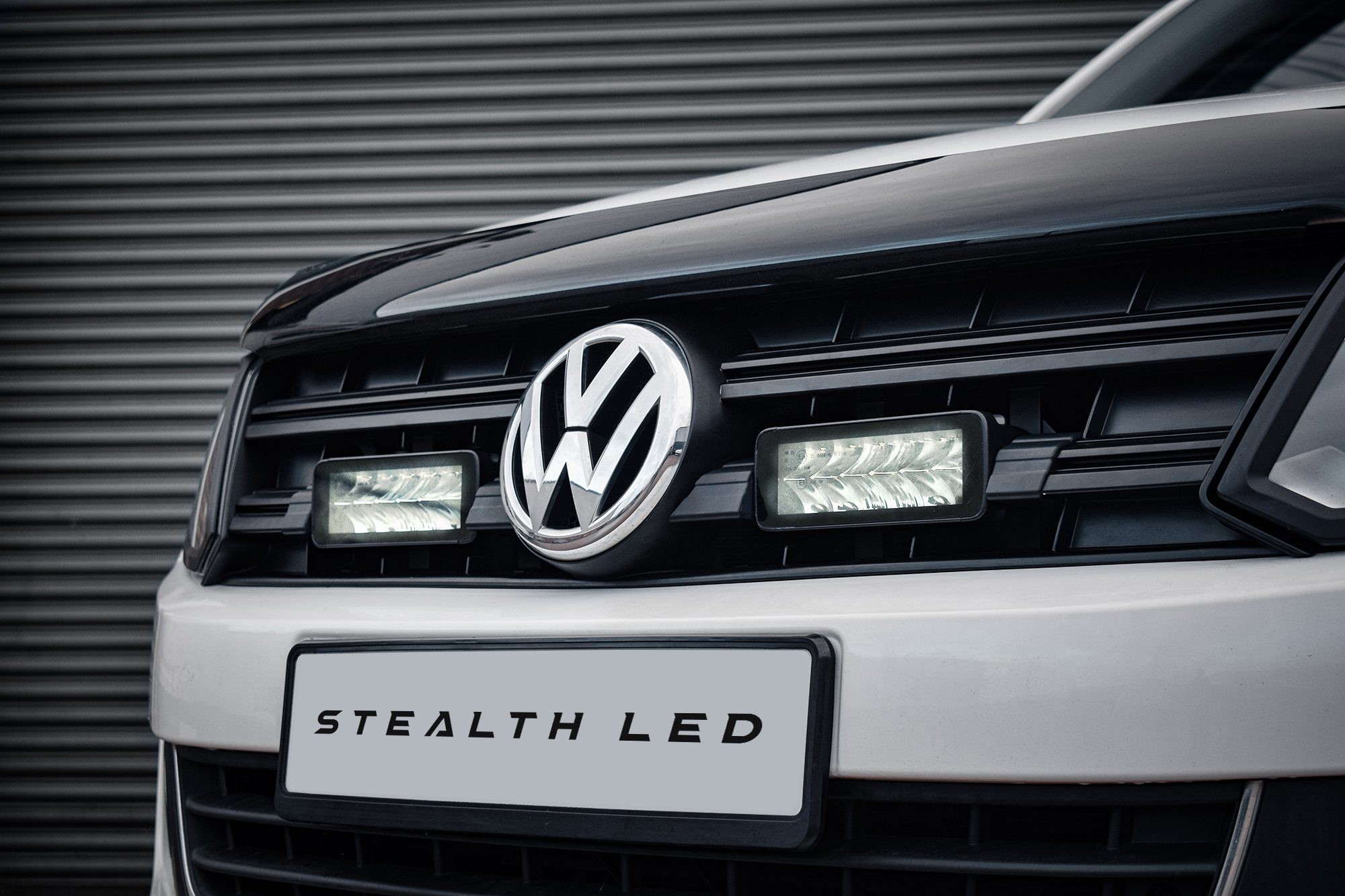 Black front grille of a Volkswagen Amarok featuring two bright, rectangular LED lights with a sleek design, accented by the VW logo and a white "Stealth LED" label below.