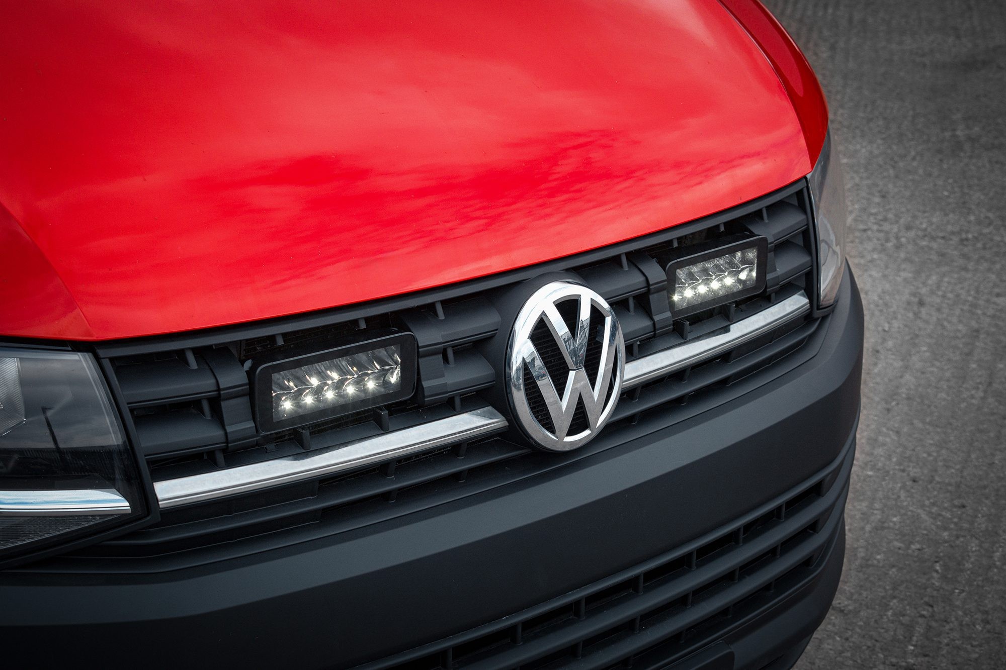 Red VW Transporter T6 with a black grille featuring twin luminous LED lights, showcasing a sleek design and the iconic VW logo prominently displayed at the center.