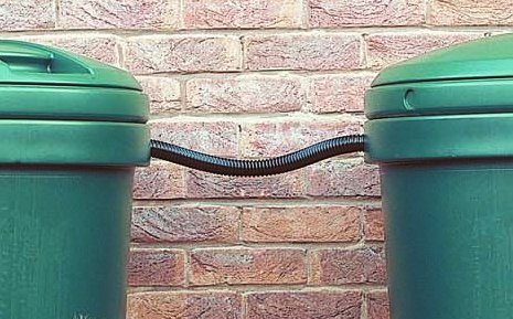 Black flexible linking hose connecting two green water butts against a brick wall, designed for efficient water collection and storage.