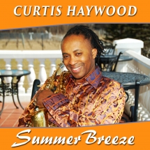 A man with long hair holds a saxophone, wearing a vibrant orange and gold patterned shirt, smiling against a backdrop of a balcony and vineyard.