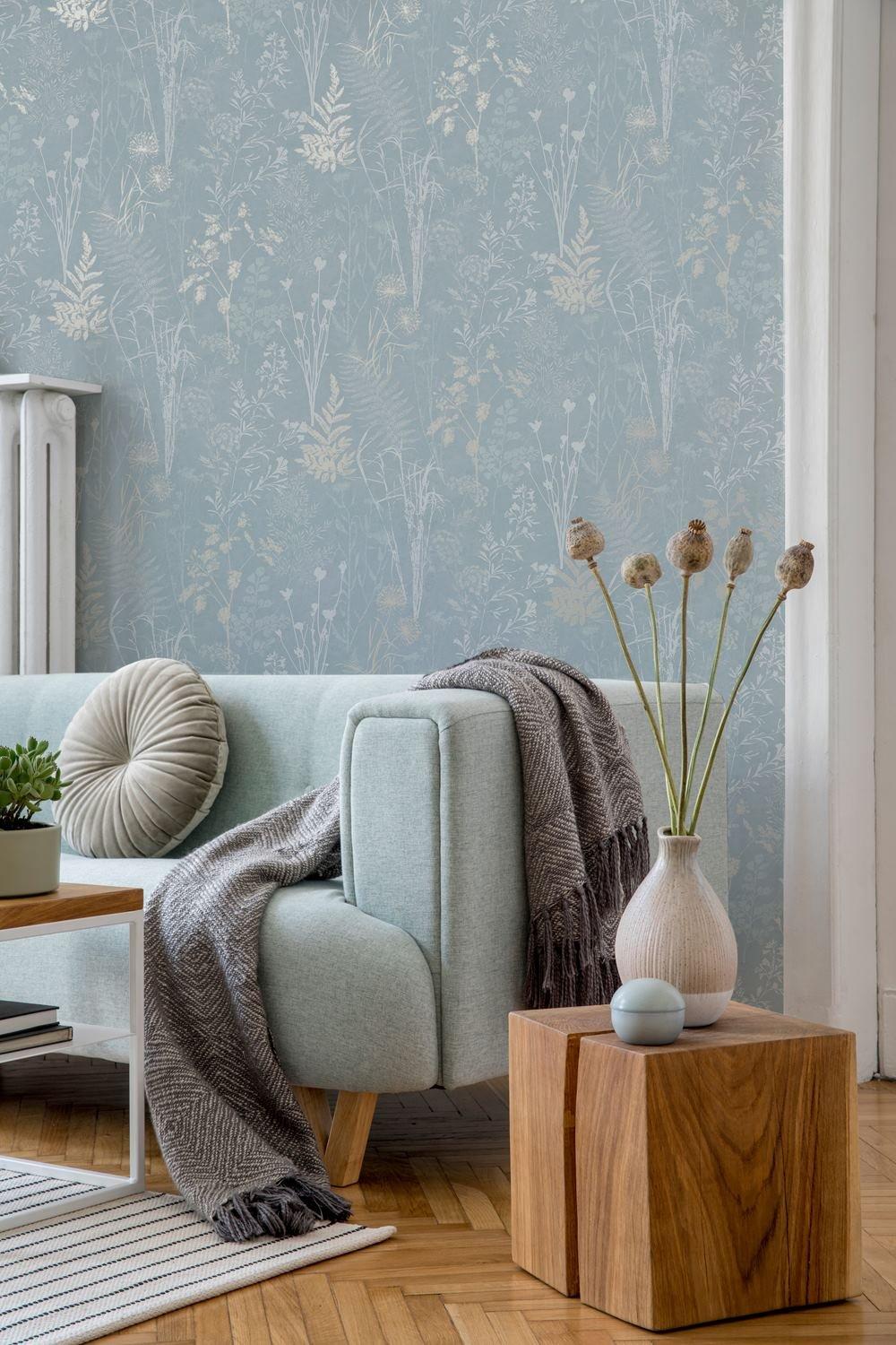 Sage metallic floral wallpaper in duck egg blue features delicate botanical patterns, complementing a light blue sofa with a cozy throw and a wooden side table.