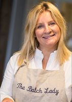 A smiling woman with long blonde hair wears a light-colored apron that reads "The Batch Lady," set against a blurred kitchen background.