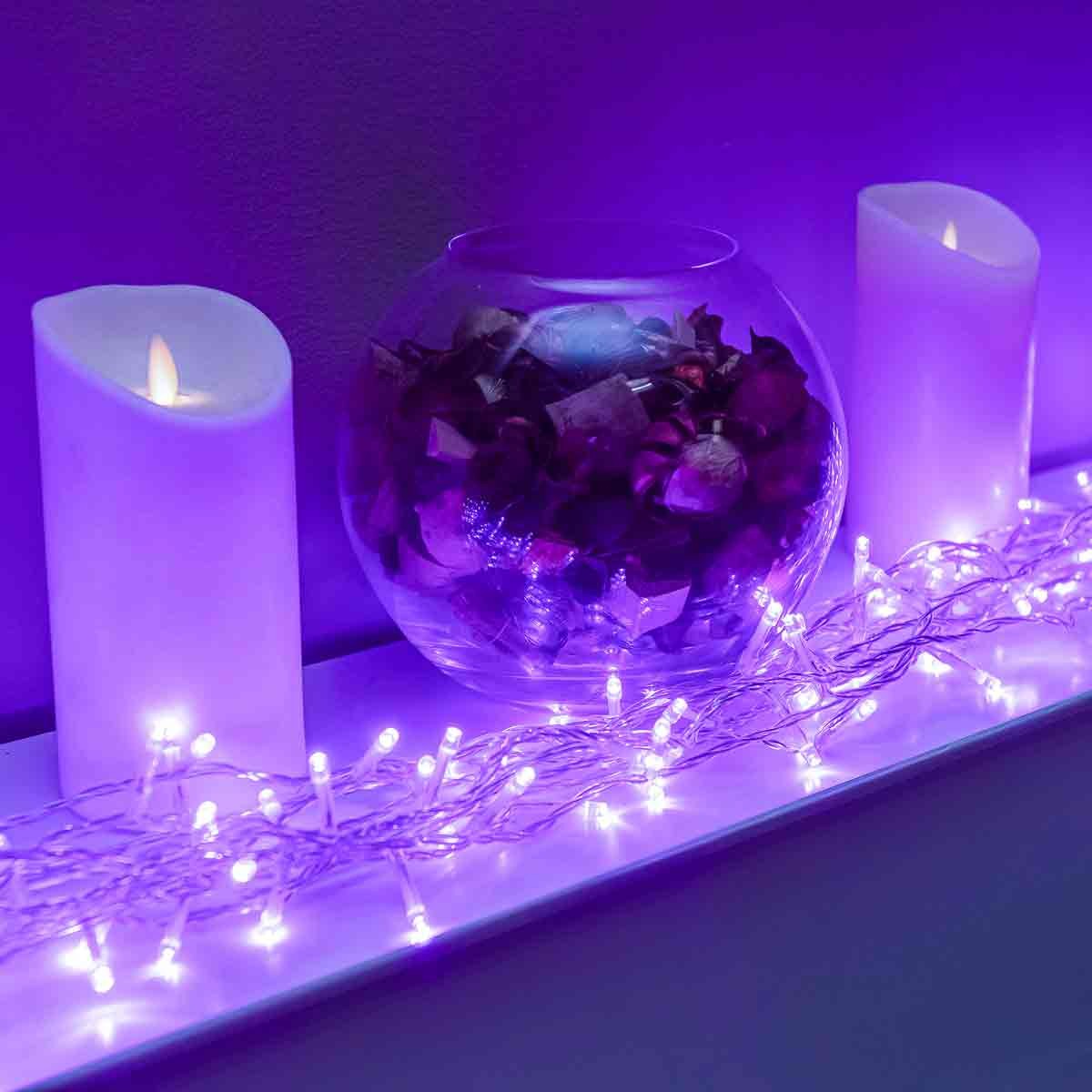120 LED purple multi-function Christmas string lights with a clear cable, displayed alongside a glass bowl of decorative items and two white flameless candles.