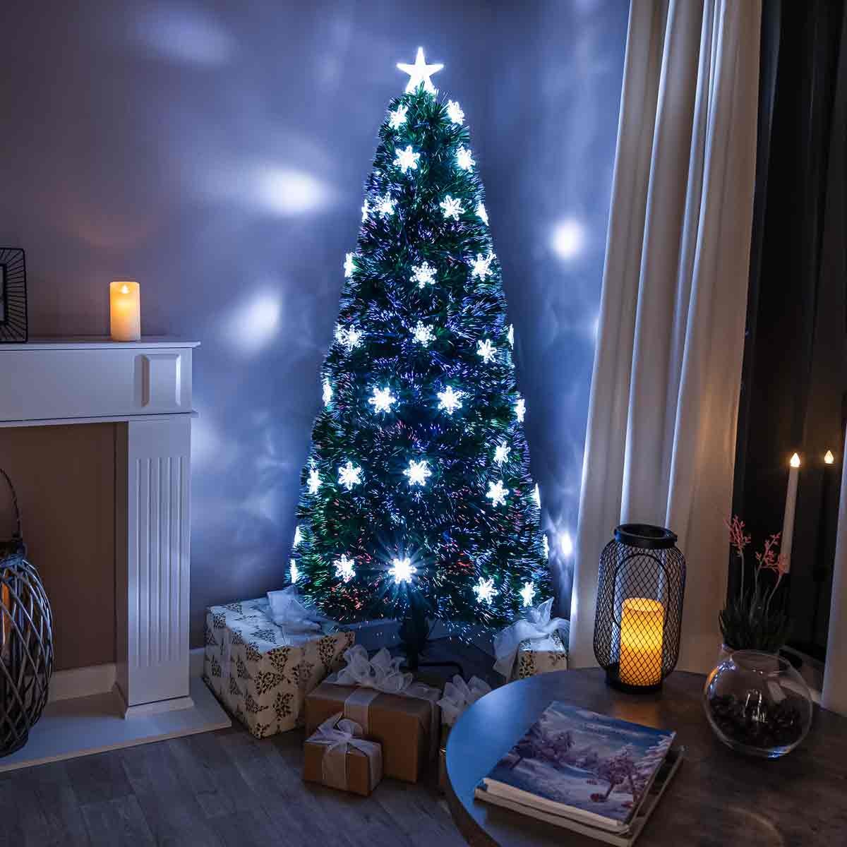 A 5-foot green fibre optic Christmas tree adorned with glowing white snowflakes and a star topper, set beside wrapped gifts and candles in a cozy room.