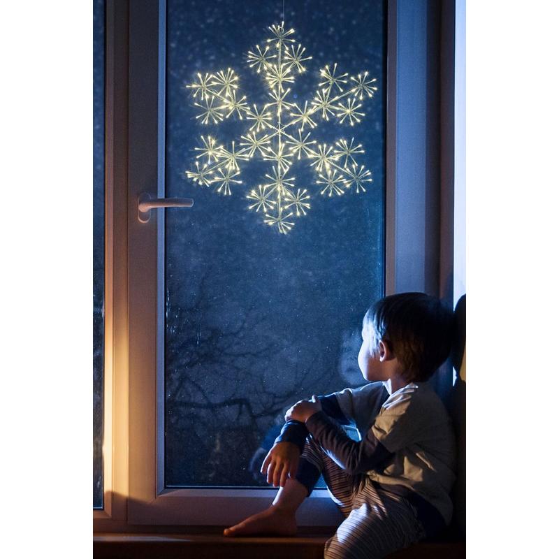 60cm warm white LED snowflake decoration with a starburst design, glowing against a dark window, while a child sits nearby, gazing at the festive display.