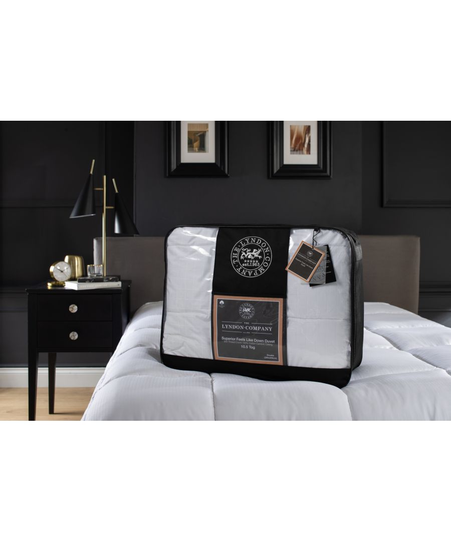 White duvet with a black and gold label, packaged in a clear plastic cover, resting on a bed with a dark headboard and a bedside table featuring a lamp and framed pictures.