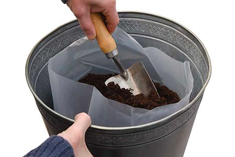 A hand holds a metal bucket with a decorative rim, lined with a plastic insert, filled with dark soil and a metal trowel resting inside.