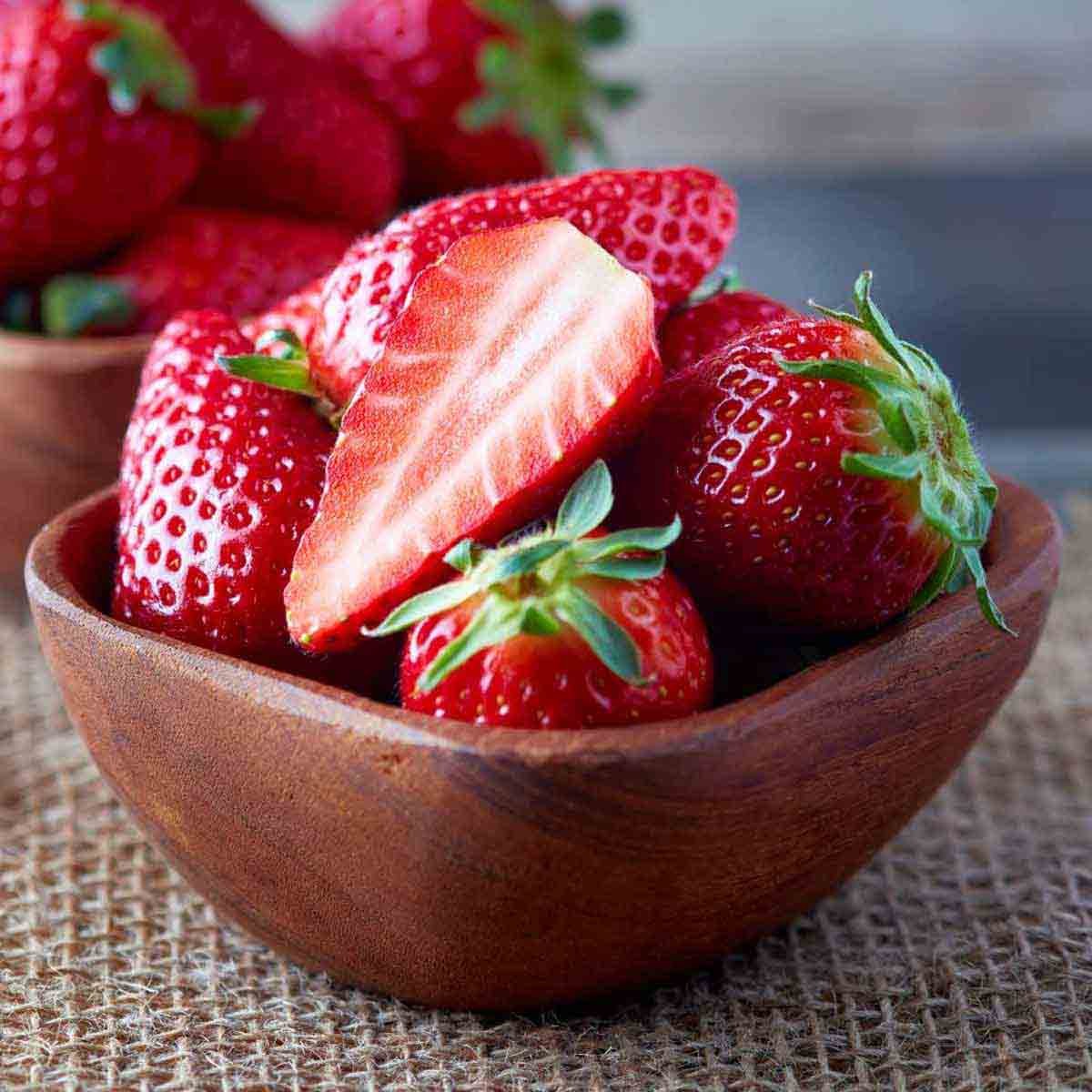 Fresh, vibrant red strawberries in a wooden bowl, with one sliced berry showcasing its juicy interior, set against a textured burlap background.